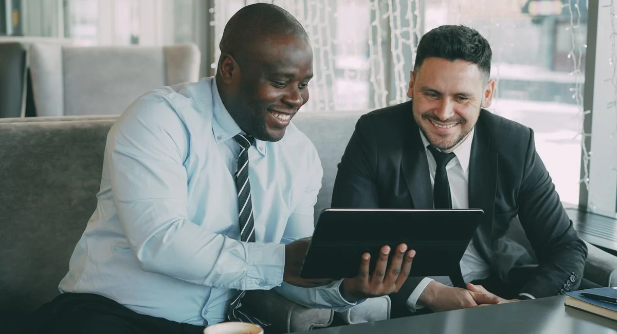 Two businessmen happily collaborating on a digital tablet in a modern cafe setting.