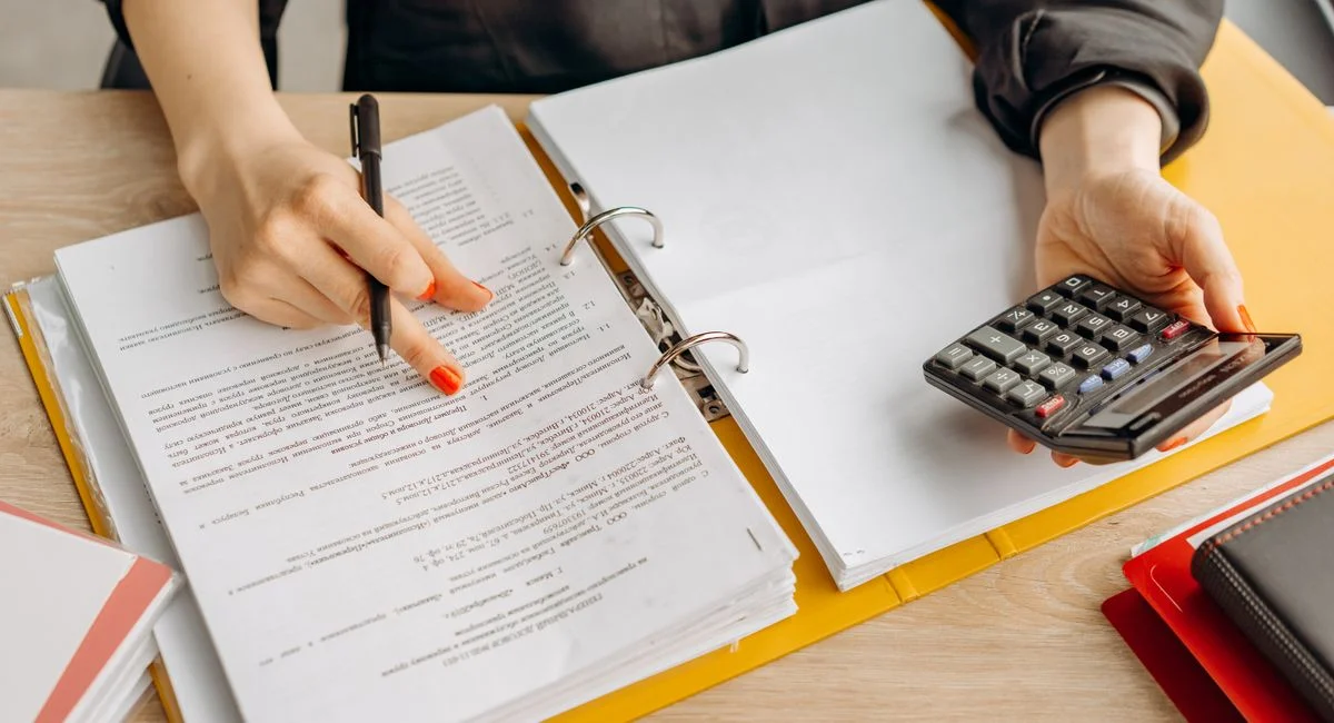 A woman reviewing documents and using a calculator on a wooden table.