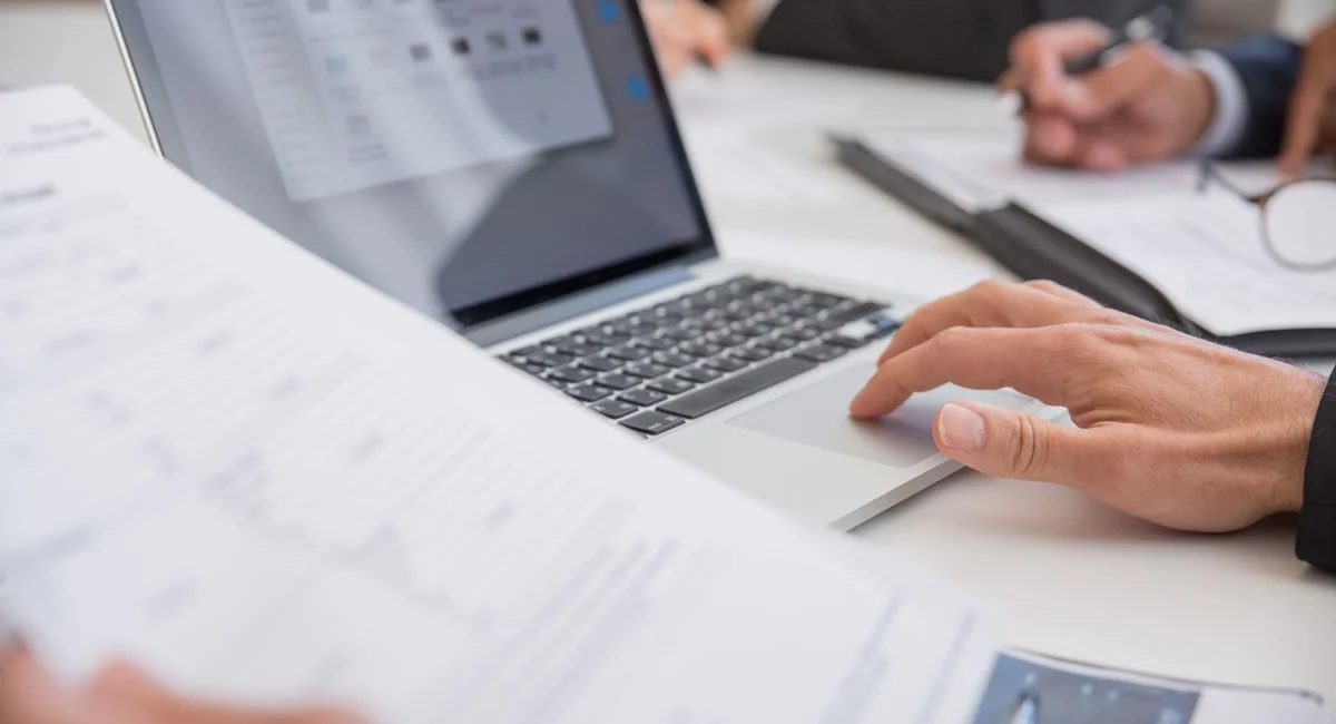 Close-up of hands working on documents and a laptop in an office setting, illustrating teamwork and productivity.