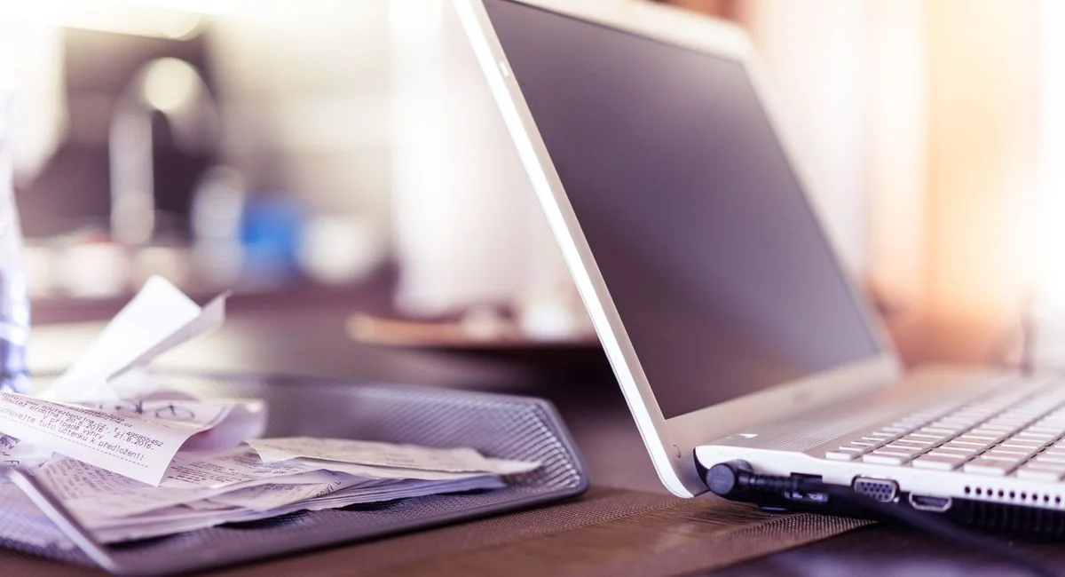 A close-up of an open laptop beside a mesh tray filled with receipts on a desk.