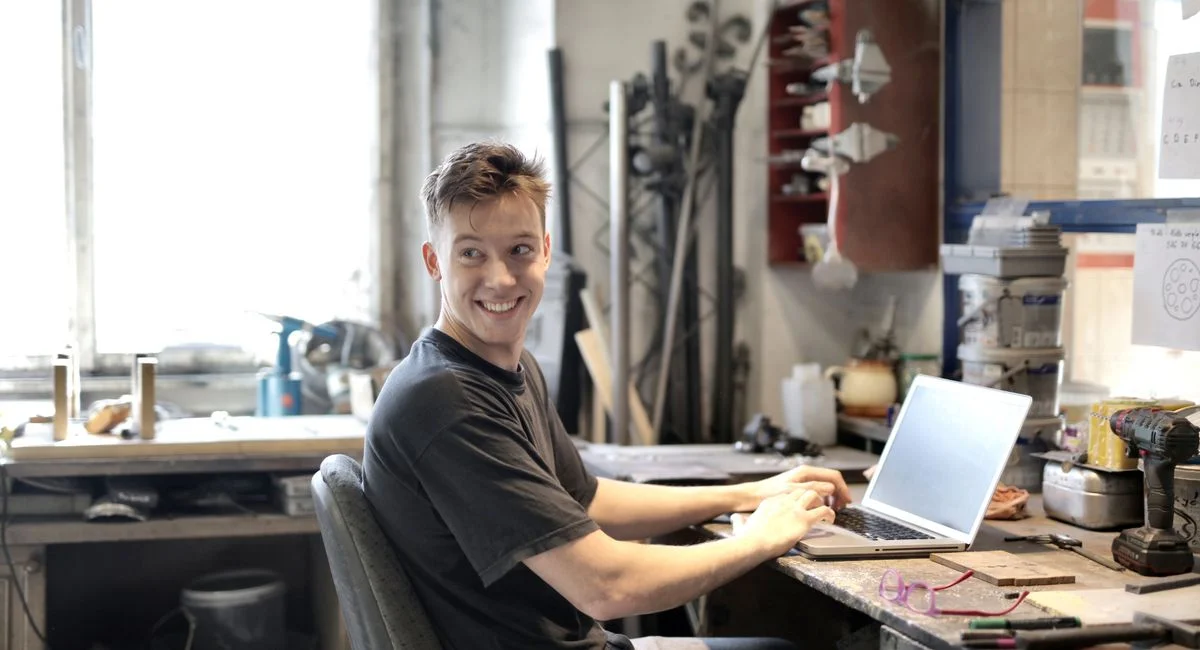 Young man smiling while using laptop in a creative workspace with tools around.