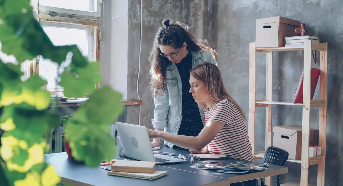 Two young women working together in a modern office with a laptop and plants.