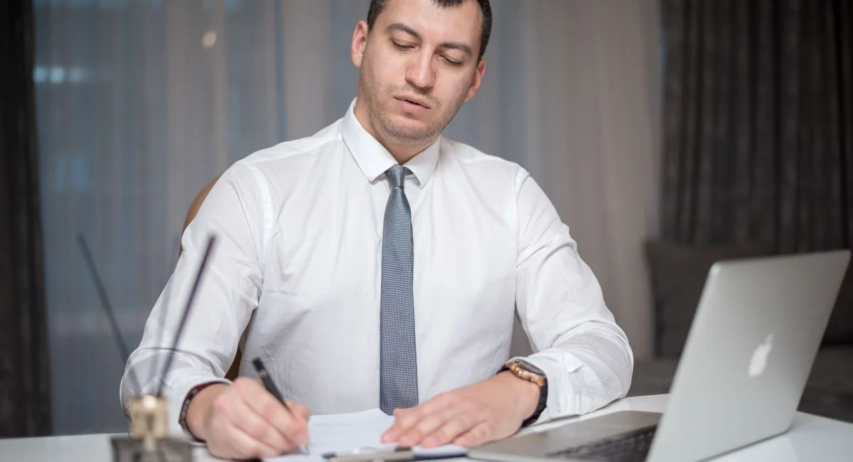 Focused businessman in office writing notes on paper with laptop at a white desk.