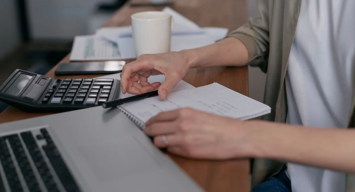 A mid-adult calculating finances with a laptop and notepad, managing budgets at home.