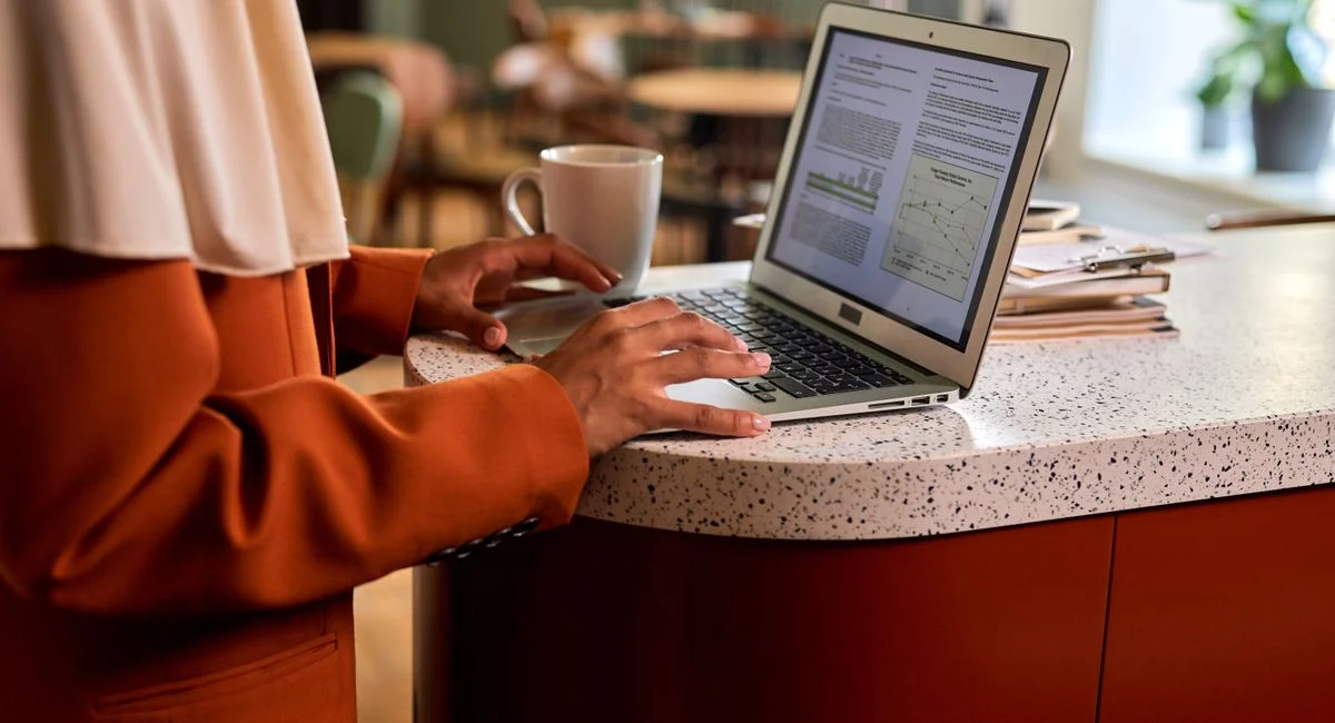 Adult woman typing on a laptop at a modern office counter, coffee beside her in a casual indoor setting.