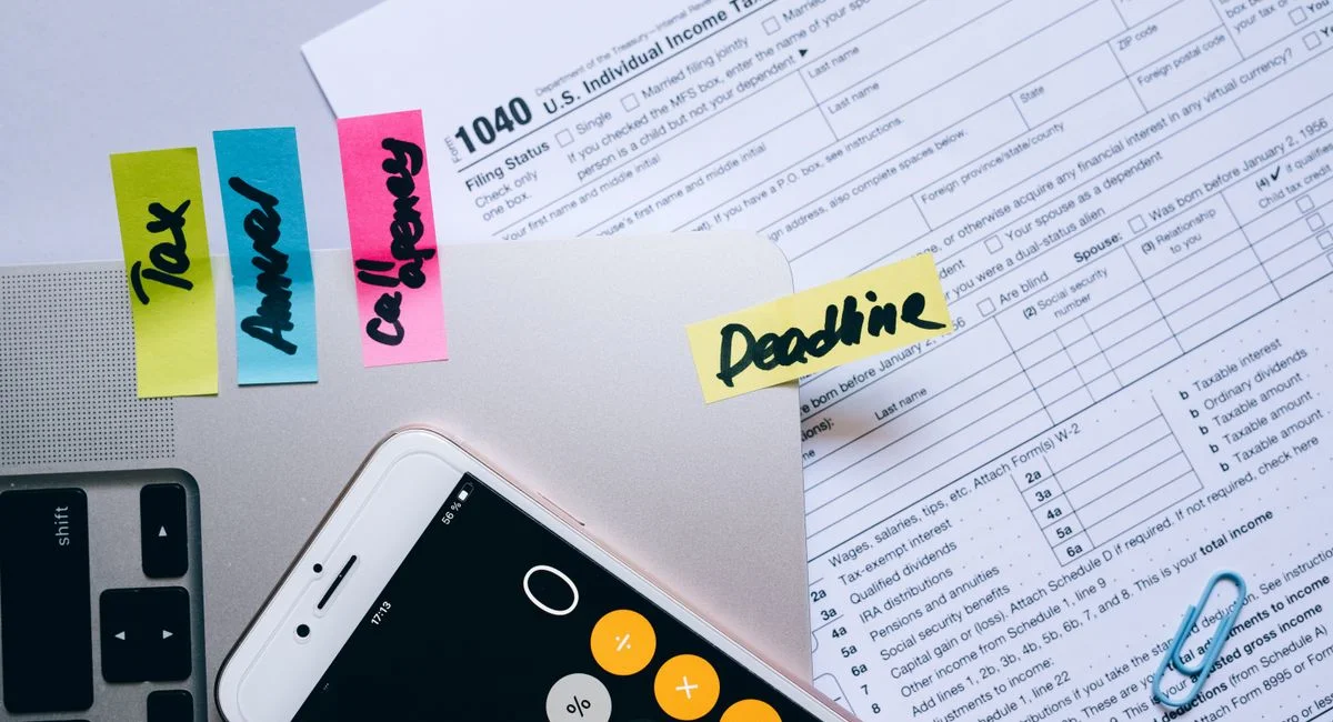 Office desk flat lay showing tax documents, calculator app on smartphone, sticky notes, and paperclips.
