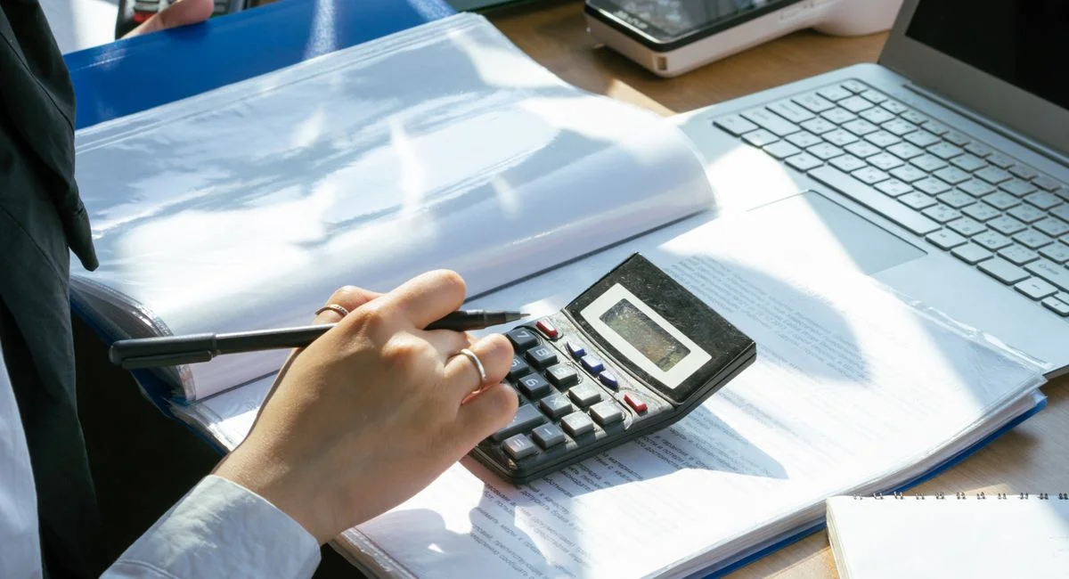 A person working on financial calculations using a calculator and laptop at an office desk.