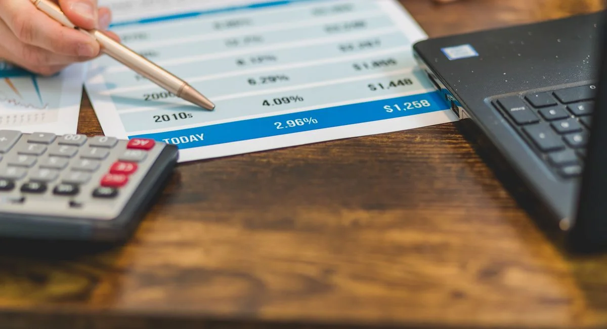 Close-up of a hand with pen analyzing financial rates on paper with a calculator and laptop nearby.