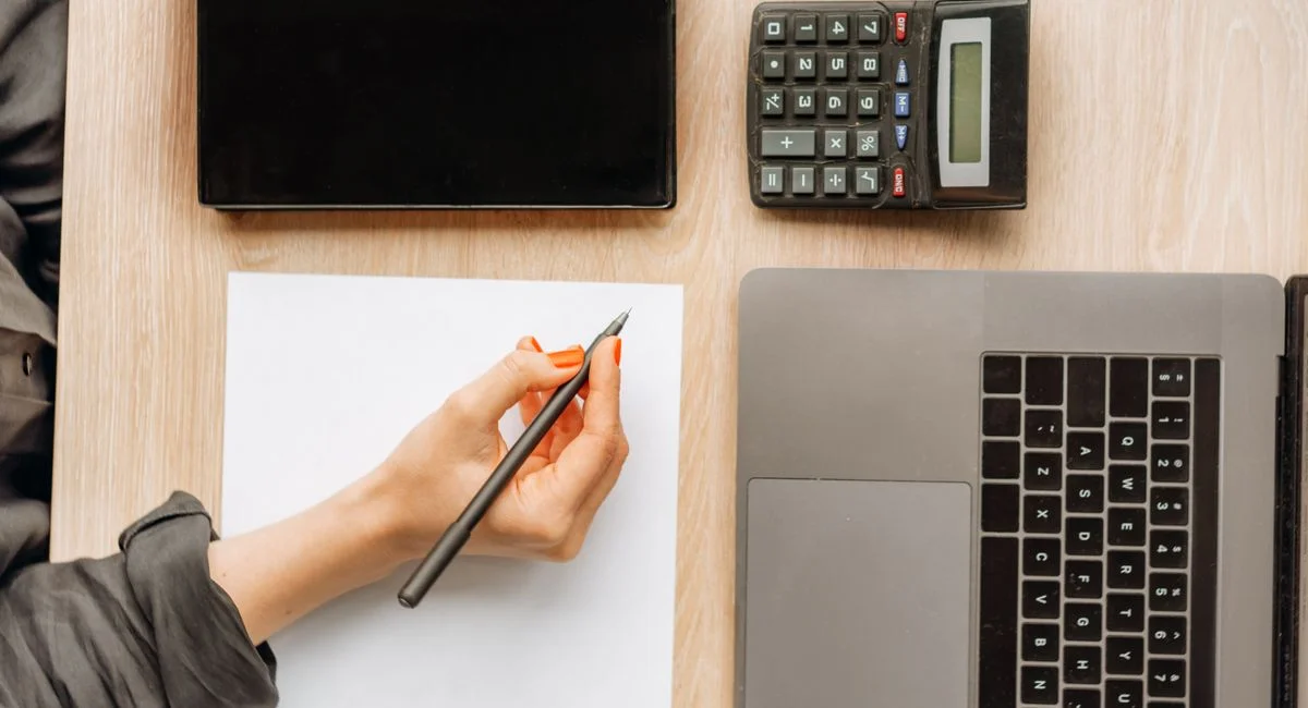 A person writing on paper surrounded by a laptop, tablet, and calculator on an office desk.