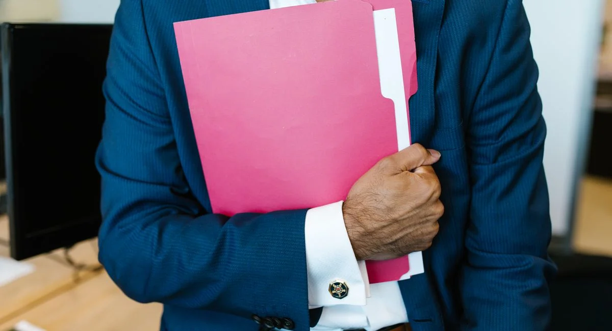 A professionally dressed man holding a pink folder in an office setting.