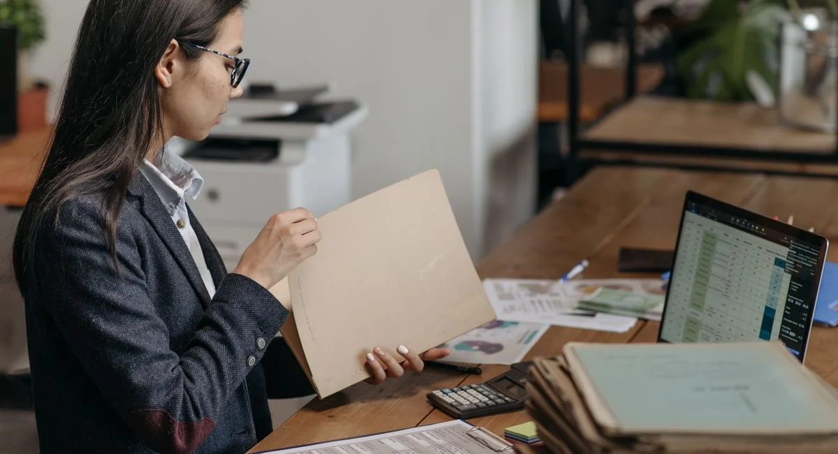 A professional woman reviewing documents at her office desk with a laptop and files.