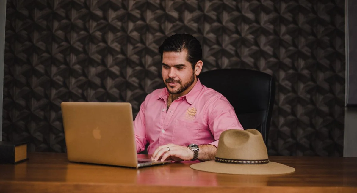 Businessman working on a laptop in an office setting, wearing a pink shirt, with a hat on the desk.
