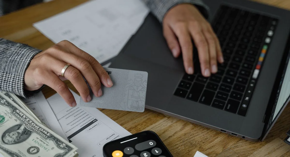 Close-up of a person using a credit card and laptop in a financial setting with cash and calculator.