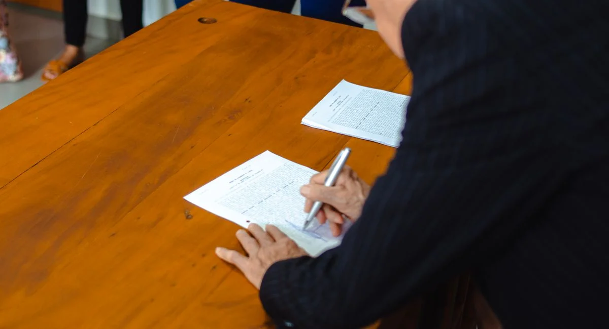 A close-up of a businessman signing official documents at a wooden desk.