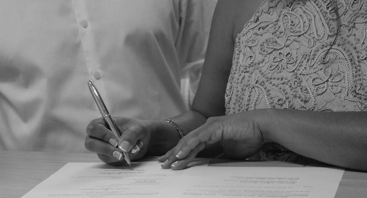Black and white image of a couple signing documents, focusing on their hands and pen.