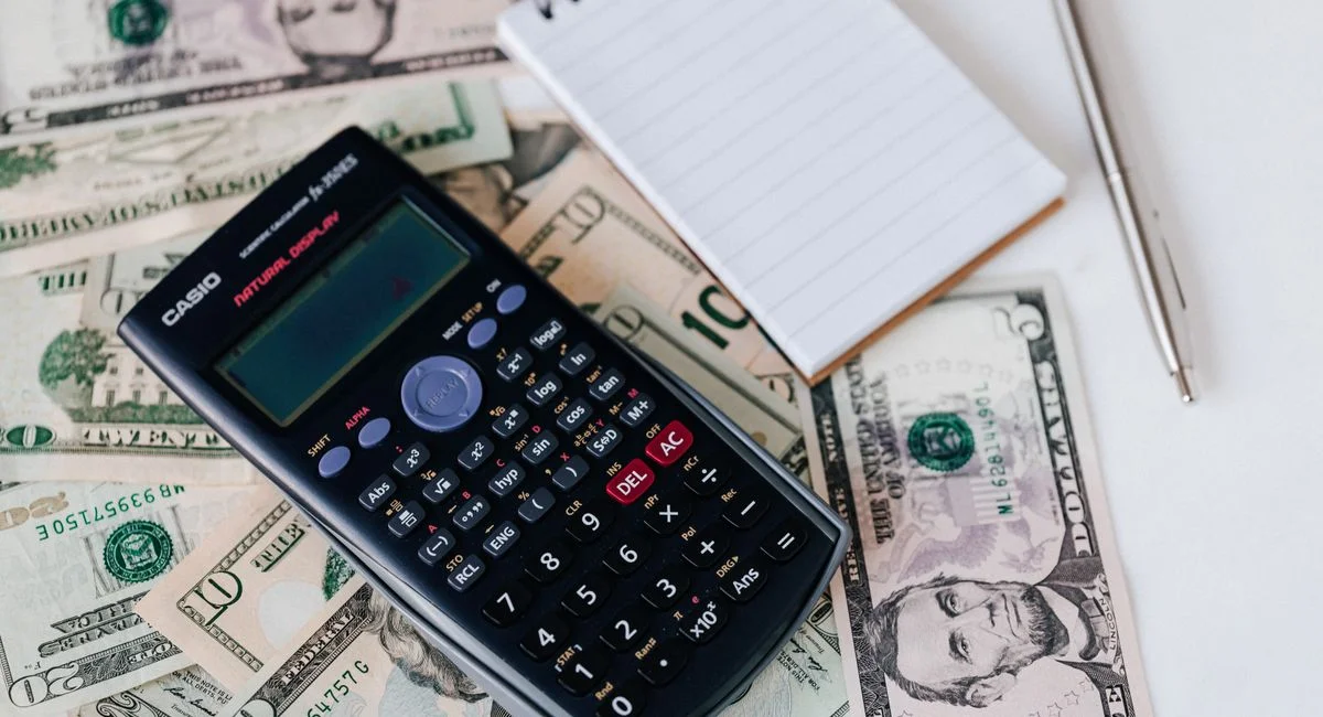 From above electronic calculator and notepad placed over United States dollar bills together with metallic pen for budget pla