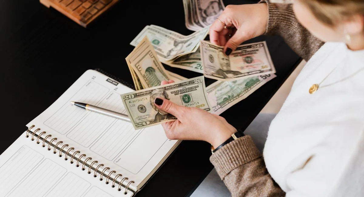Woman organizing finances, counting dollar bills at desk with open notebook.