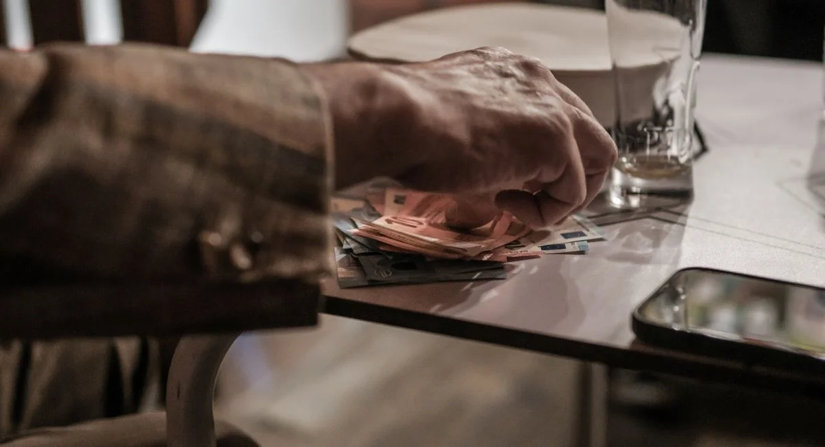 Hand counting euro banknotes on a table beside a phone and empty glass.