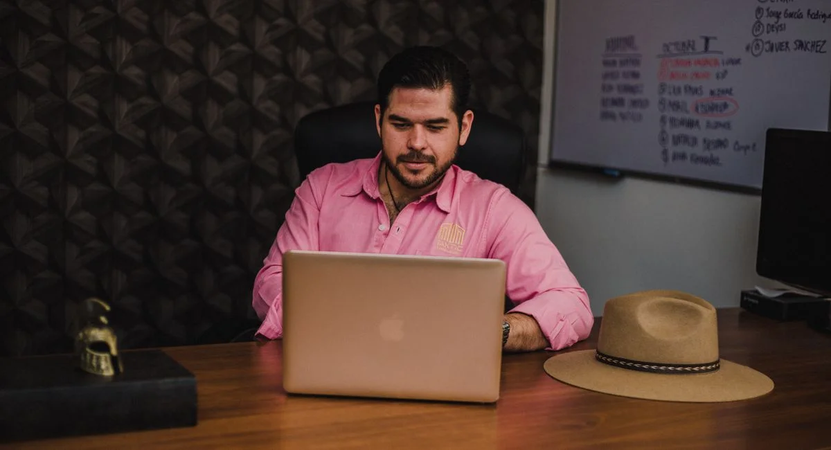 Man in pink shirt working on laptop at a wooden desk in office.